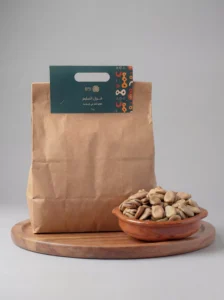 كيس ورقي بني صديق للبيئة لمنتج "فول السليم" من علامة سدرة التجارية، مثبت عليه ملصق أخضر داكن يحمل شعاراً ذهبياً وعبارة "طعم البلد في كل وجبة". يظهر بجانب الكيس طبق فخاري تقليدي ممتلئ بحبات الفول السوداني العريضة والنقية، موضوعة على قاعدة خشبية دائرية بخلفية رمادية أنيقة.