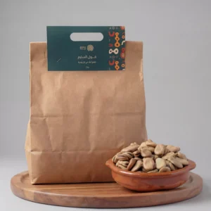 كيس ورقي بني صديق للبيئة لمنتج "فول السليم" من علامة سدرة التجارية، مثبت عليه ملصق أخضر داكن يحمل شعاراً ذهبياً وعبارة "طعم البلد في كل وجبة". يظهر بجانب الكيس طبق فخاري تقليدي ممتلئ بحبات الفول السوداني العريضة والنقية، موضوعة على قاعدة خشبية دائرية بخلفية رمادية أنيقة.